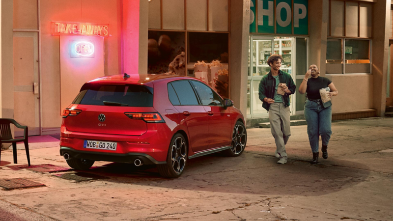 Rückansicht eines roten VW Golf GTI, der vor einem Kiosk steht. Ein Mann und eine Frau mit Snacks in der Hand kommen auf einen zu.