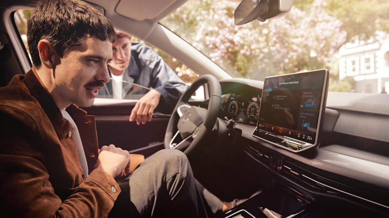 Un homme est assis sur le siège conducteur et regarde le système multimédia. Un deuxième homme se tient à l'extérieur de la voiture et se penche légèrement à l'intérieur par la fenêtre ouverte.