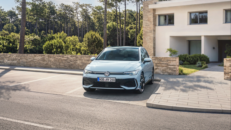 A light-blue VW Golf hybrid in a parking lot in front of a residential house by the forest, front view.