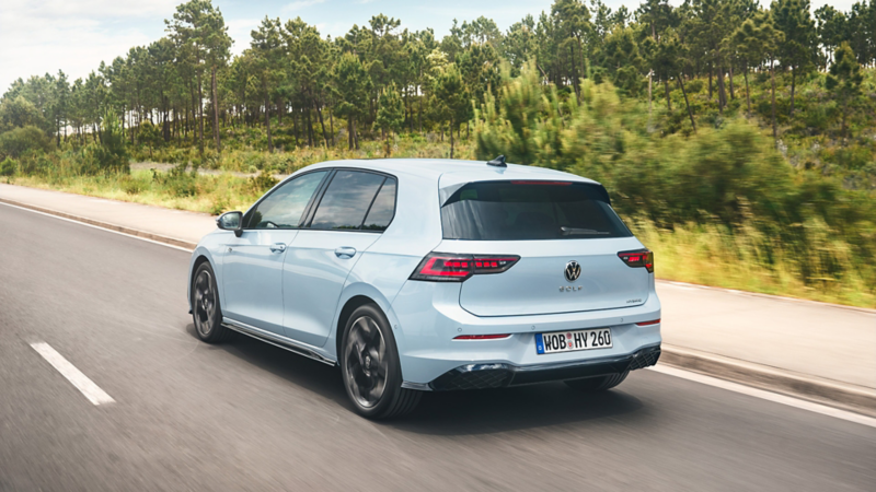 Ein hellblauer VW Golf Hybrid fährt auf einer Straße an einem Wald vorbei.