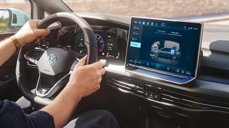 Wielofunkcyjna kierownica i system infotainment w hybrydzie VW Golf, osoba prowadzi pojazd.