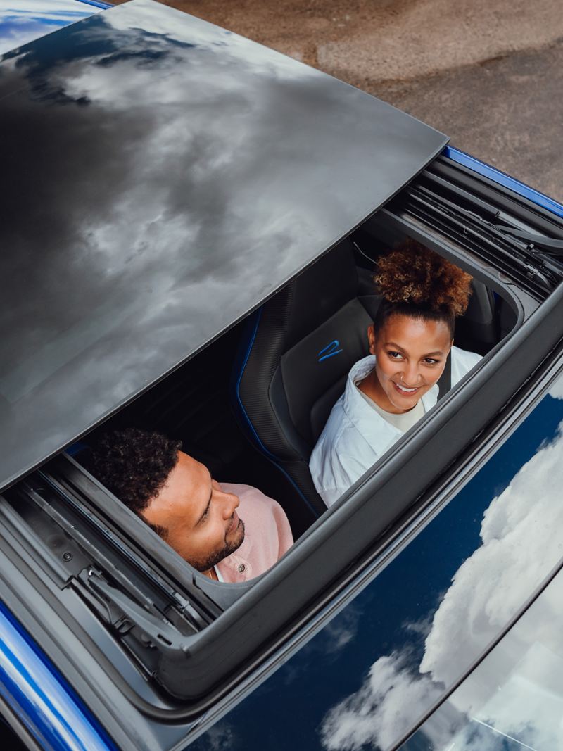 A couple sits in the VW and holds hands, while the woman looks through the open sunroof into the sky