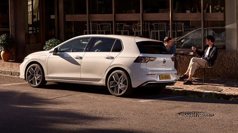 Une Golf blanche est garée au bord de la route. Deux hommes sont assis sur le trottoir et boivent du café.