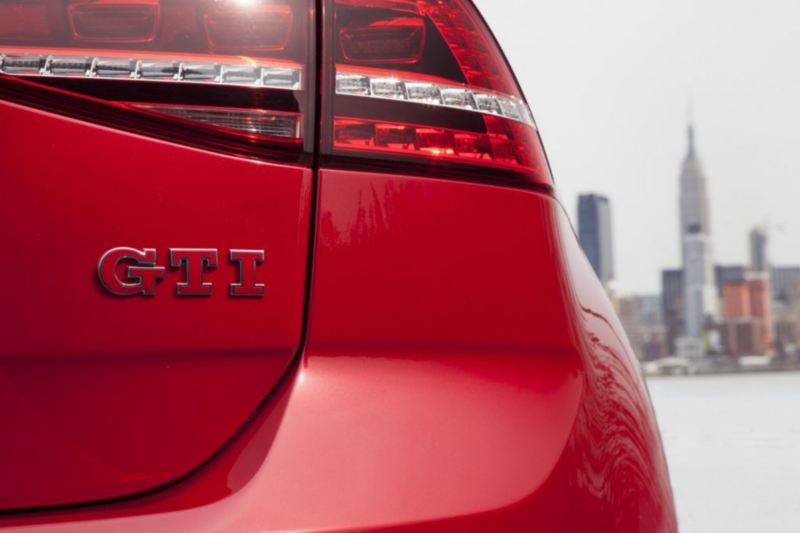 Se muestra un Volkswagen Golf GTI rojo desde atrás, con el emblema en primer plano contra el horizonte de la ciudad.