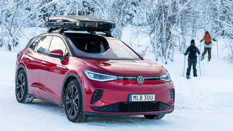 En röd ID.4 GTX med svart takbox står parkerad i vinterlandskap, två personer på skidor syns i bakgrunden