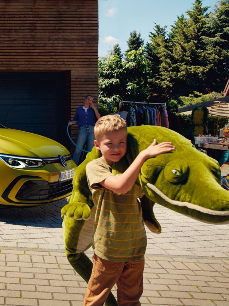 VW Golf Variant in Gelb parkt seitlich vor einer Garage, im Vordergrund ein Junge mit riesigem Kuscheltier.
