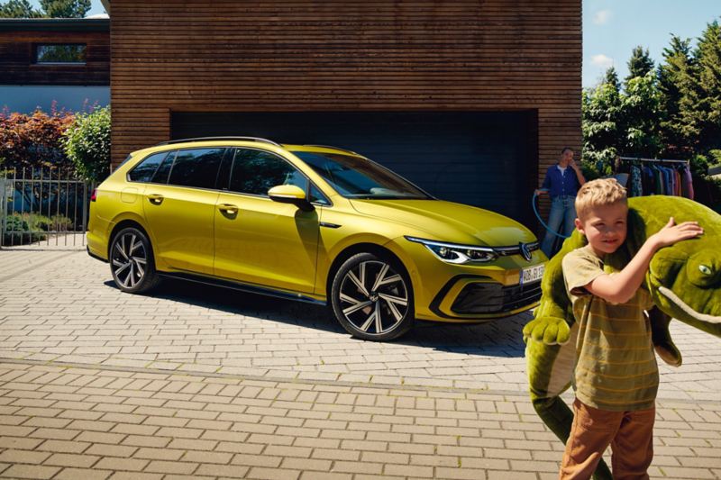 En gul VW Golf Variant holder parkeret ved siden af en garage, i forgrunden ses en dreng med et kæmpe tøjdyr.