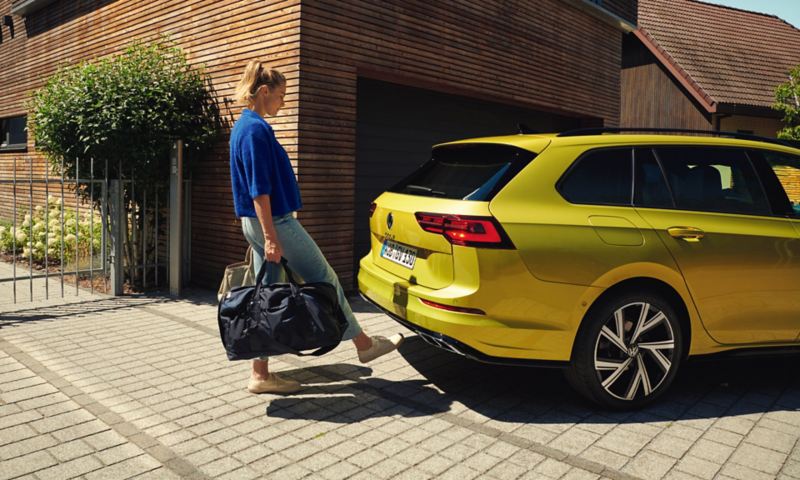 Frau mit Taschen in den Händen öffnet Heckklappe des VW Golf Variant in Gelb mit dem Fuss über das optionale «Easy Open & Close".