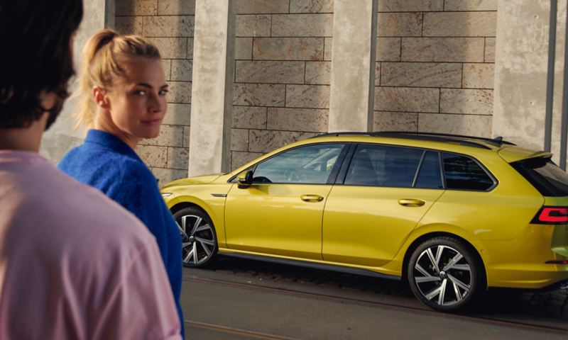 VW Golf Variant in Gelb mit optionalen getönten Scheiben von der Seite, Mann und Frau gehen darauf zu.