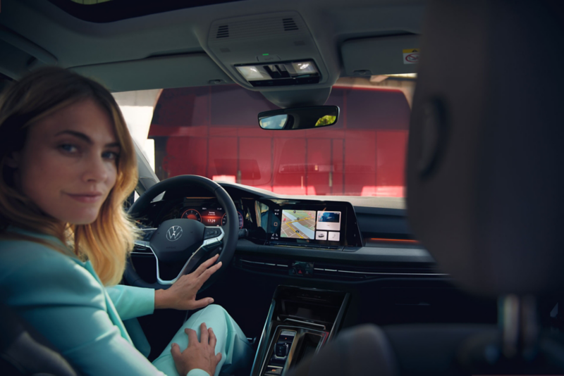 Vue depuis les sièges arrières d'une Golf, vers l'avant, une femme assise sur le siège conducteur se retourne et sourit.