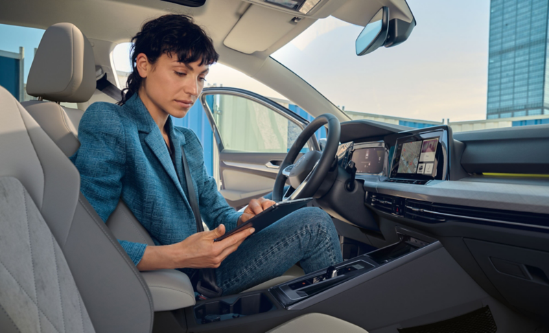 Blick vom Beifahrersitz auf eine Frau auf dem Fahrersitz im VW Golf, helles Interieur. Sie nutzt den integrierten WLAN-Hotspot über ein Tablet.
