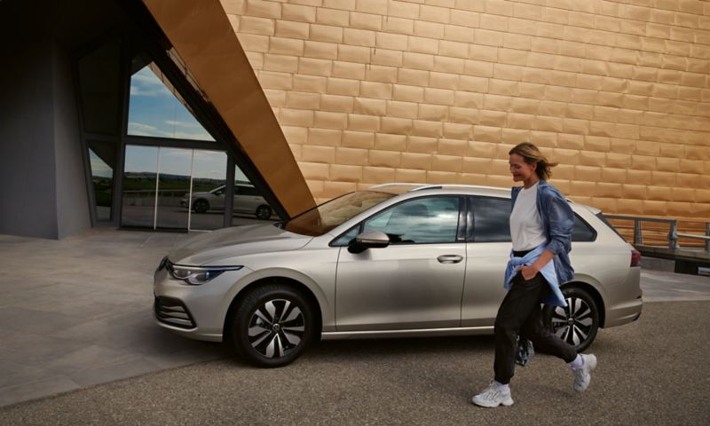 VW Golf Kombi MOVE in silber parkt vor einem futuristischen Gebäude, Blick auf Seite und Felgen, eine Frau geht daran vorbei