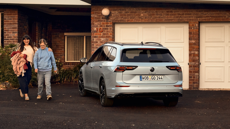 En vit VW Golf sportscombi parkerad framför en garageport, en kvinna och en ung tjej går bredivd bilen 