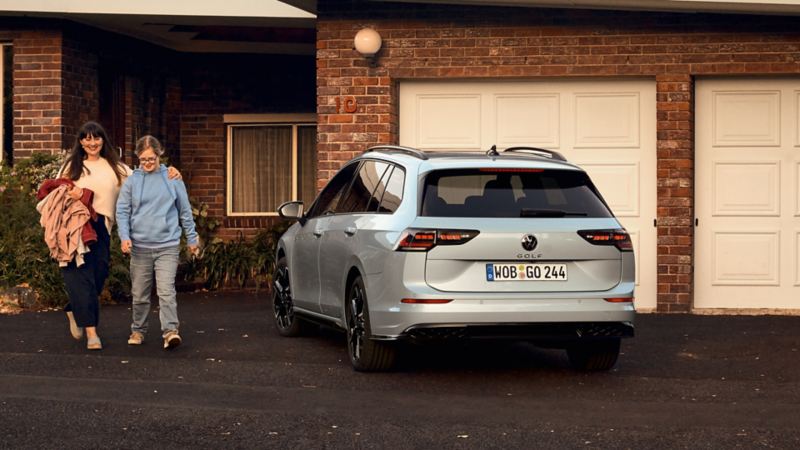 Une VW Golf SW garée devant la porte de garage d'une maison. Une femme et son enfant sortent de la maison.