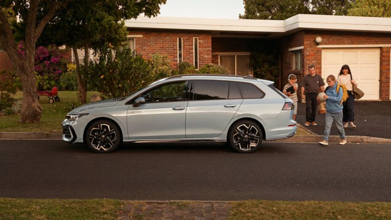 Una famiglia con i bagagli si avvicina a una VW Golf Estate bianca parcheggiata davanti a una casa.