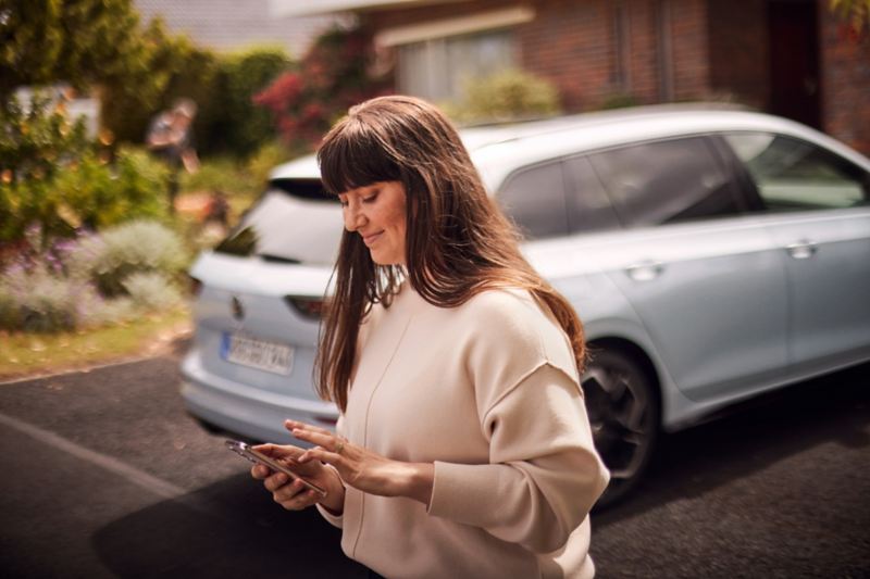 Eine Frau blickt außerhalb ihres weißen VW Golf auf ihr Smartphone.