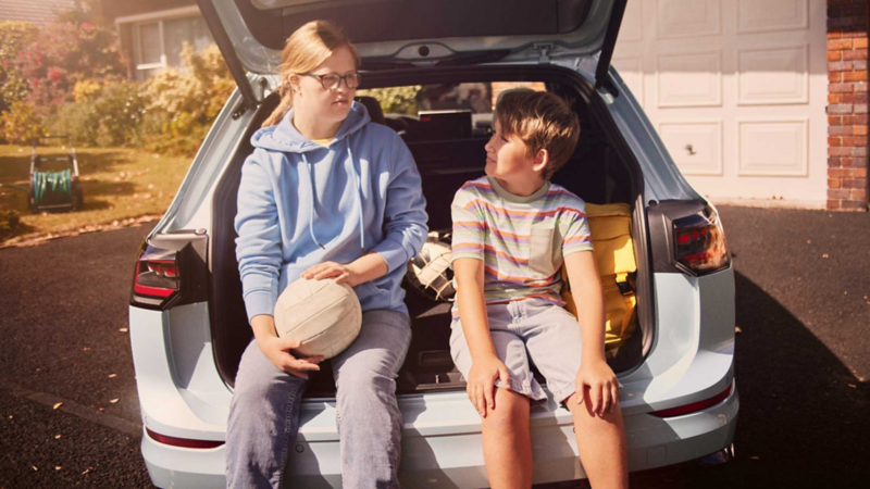 Junge und Mädchen sitzen im offenen Kofferraum eines VW Golf Variant in einer Auffahrt.