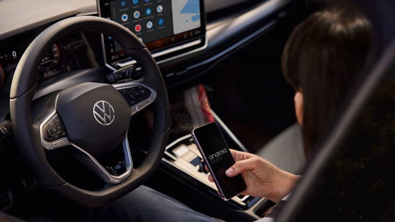 Une VW Golf GTE blanche est en train de se recharger à une station de recharge. Un homme est en train de travailler sur son smartphone au premier plan.