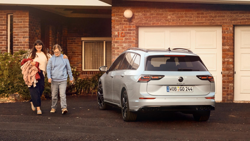 Weißer VW Golf Estate parkt vor der Garagentür eines Hauses. Eine Frau und ihr Kind gehen aus dem Haus raus.