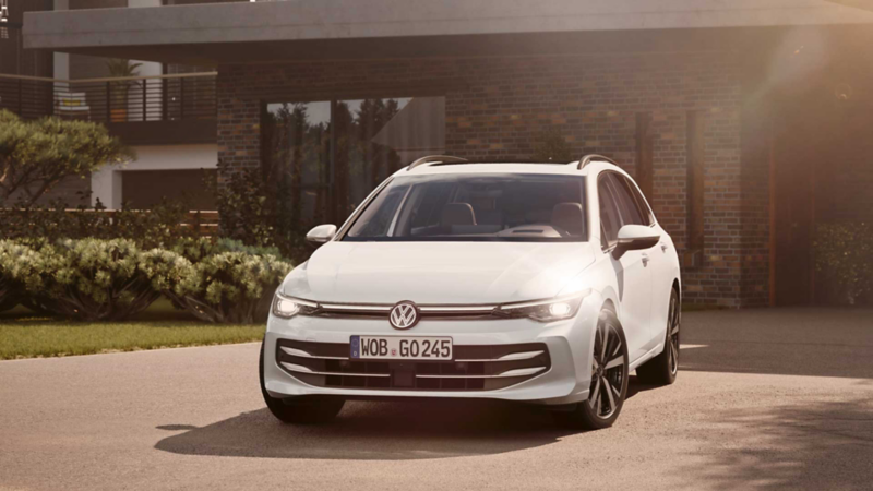 Ein weißer VW Golf Variant parkt in der Auffahrt eines Bungalows mit aktivem Tagfahrlicht.