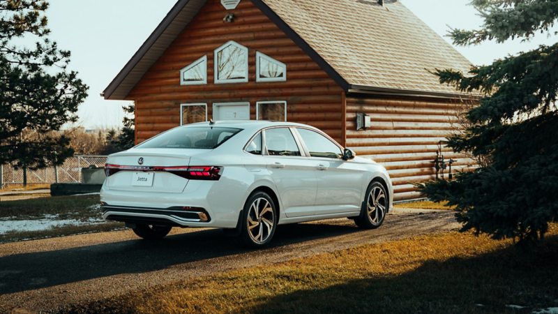 Rear view of a white Volkswagen Jetta parked in front of a log cabin.