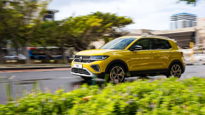 A man puts a large picnic basket in the trunk of a yellow VW T-Cross that is parked on a road in a hilly landscape.