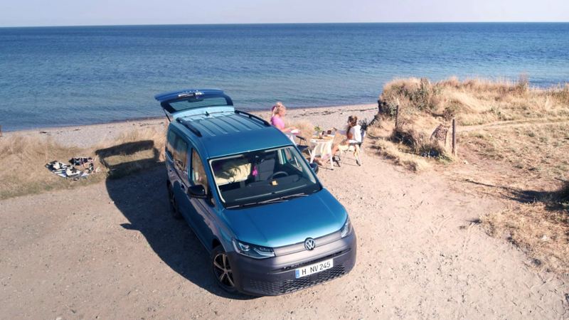 Volkswagen Caddy California parked at beach front with boot open, with people having a picnic by the beach.