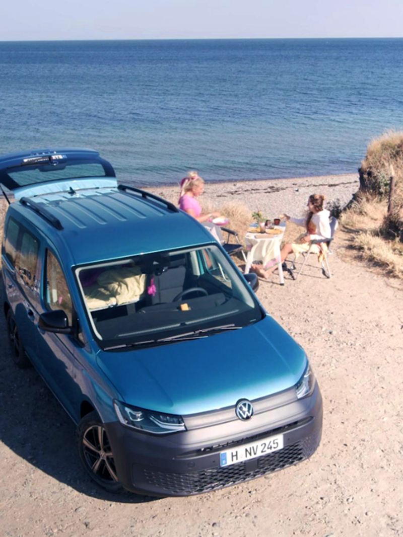 Volkswagen Caddy California parked at beach front with boot open, with people having a picnic by the beach.