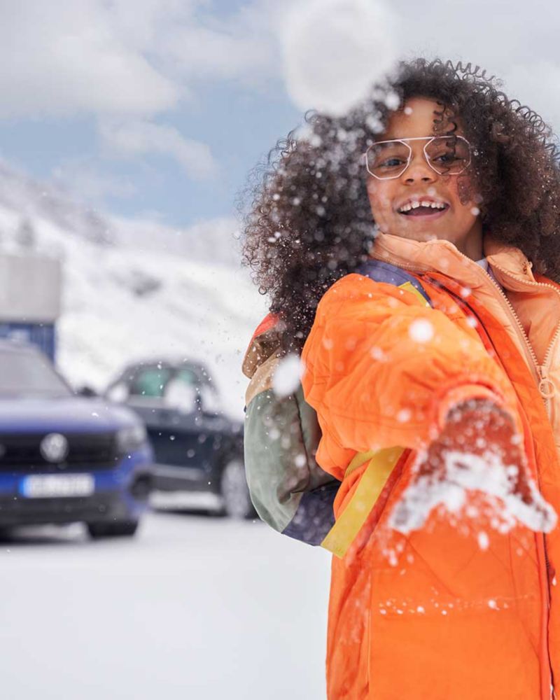 Primo piano di una bambina sorridente mentre gioca con la neve e dietro di lei parcheggiate delle auto Volkswagen.