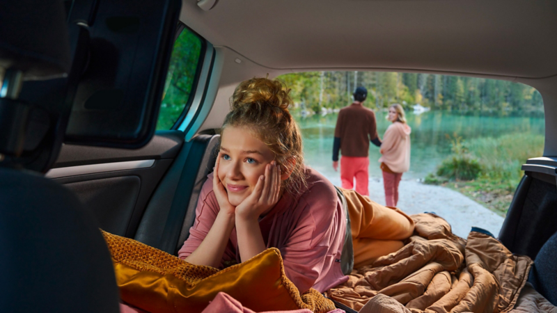 Teenager lying in the luggage compartment of a VW car, her parents can be seen through the open luggage compartment lid