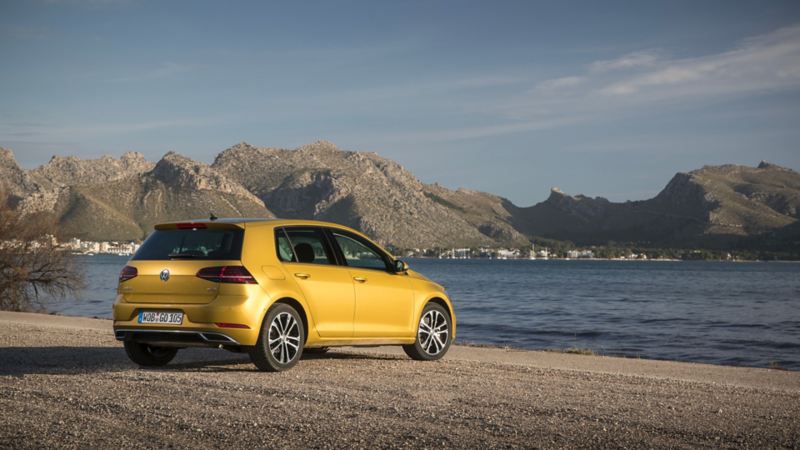 Volkswagen Golf 7 de couleur dorée, garée sur un chemin de terre au bord de la mer, avec un paysage de montagne en arrière-plan