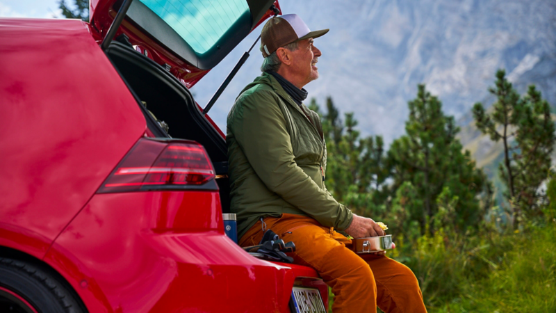 Man with a food container in his hand sits on luggage compartment lid of a VW Golf 7 and looks out into nature