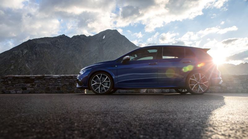 The new Golf R stands on a mountain road. In the background mountains, clouds and sun.