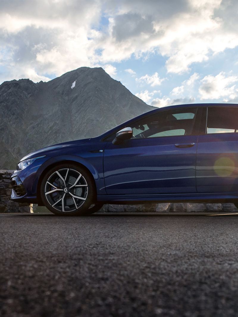 The new Golf R stands on a mountain road. In the background mountains, clouds and sun.