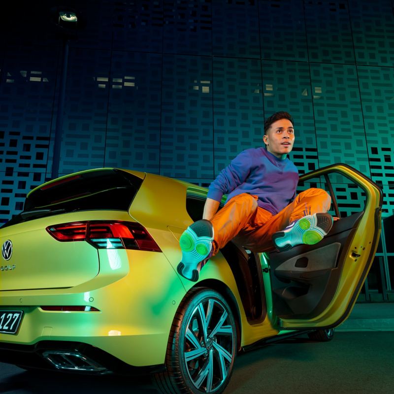 Man jumping high with pomelo yellow Volkswagen Golf parked in background.