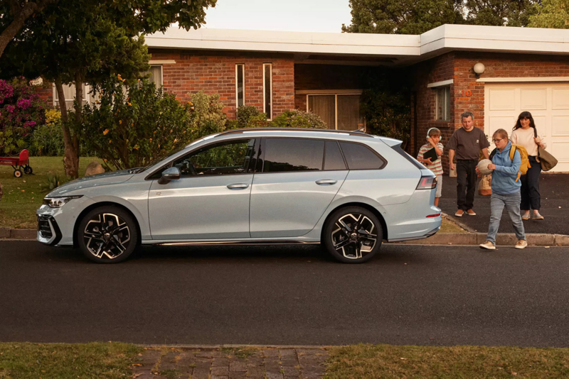 Zijaanzicht van een lichtblauwe Volkswagen Golf Variant die geparkeerd staat voor een woning. Een gezin loopt vanuit de woning richting de auto