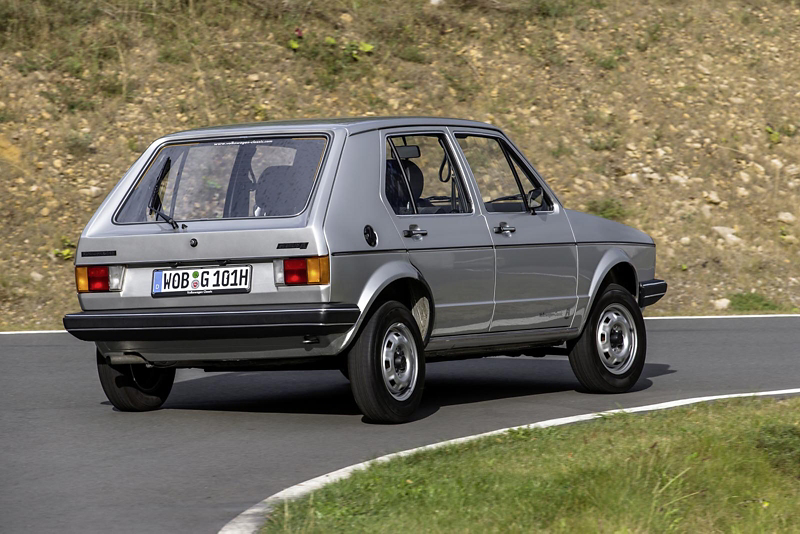 Classic grey Volkswagen Golf Mk1 drives along a winding road, showcasing the rear view and iconic hatchback design.