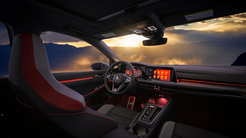 L’intérieur de la Golf GTI doté du système audio Harman / KardonMD, d’un volant chauffant et d’un tableau de bord avec des témoins DEL rouges devant les montages, le soleil se couchant en arrière-plan.