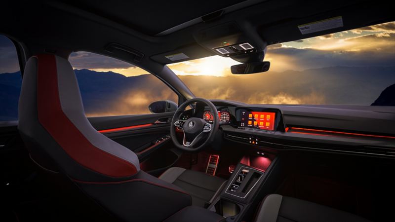 Golf GTI interior featuring Harman / Kardon® audio system, heated steering wheel and a dashboard with red LED light in front of mountains with the sun setting in the background.