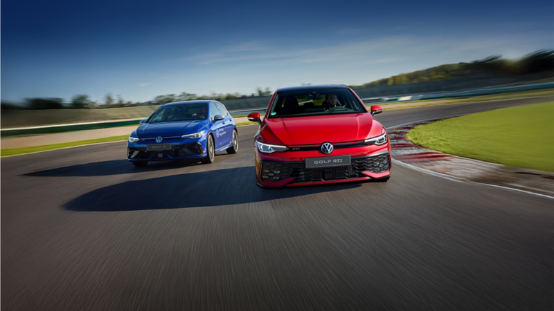 A red Volkswagen Golf GTI leads a blue Volkswagen Golf R on a race track under a clear blue sky.