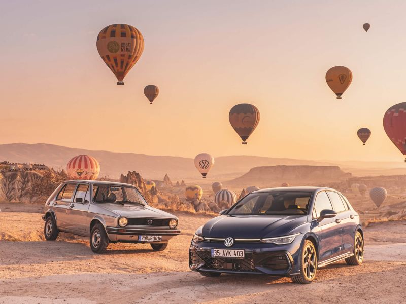 an old and a new version of the Volkswagen Golf in the desert surrounded by hot air balloons