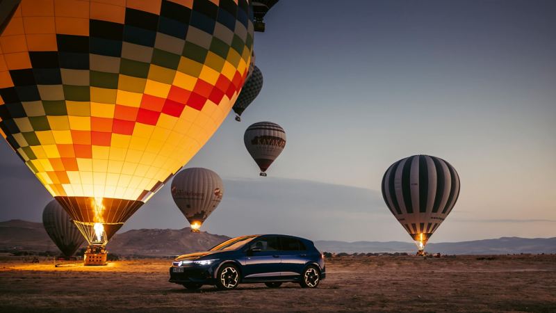 a blue Golf driving in the desert with hot air balloons floating in the background