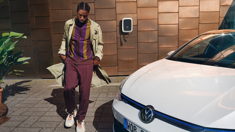 Woman strolls past a fruit stall on a market, a white VW Golf GTE is parked at the roadside.