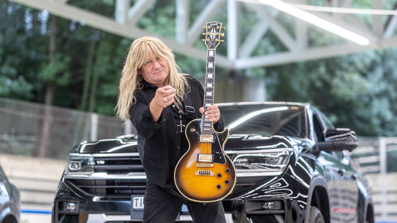 Leo Leoni steht vor seinem VW und hält eine Gitarre in der Hand.