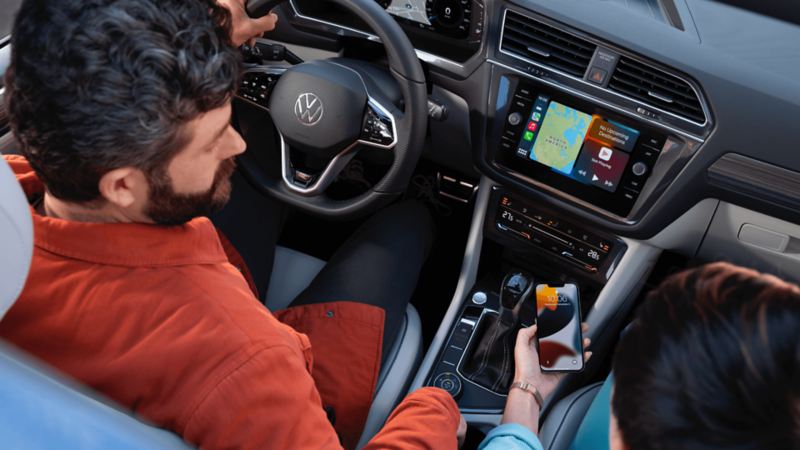 An overhead view of the interior front two seats of a 2022 Tiguan displaying the front console, steering wheel, and a man and a woman seated looking at the cellphone in the woman’s hand.