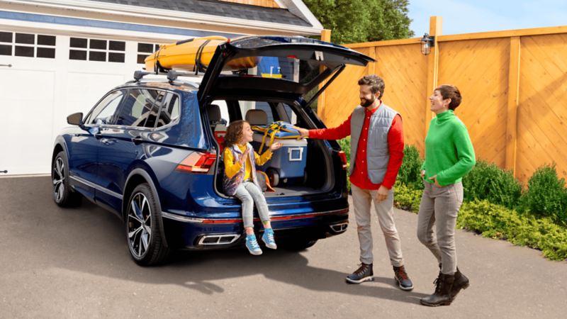 A 2022 Tiguan sits in a driveway with the tailgate open and a young girl sitting inside talking to her parents in front of her. 