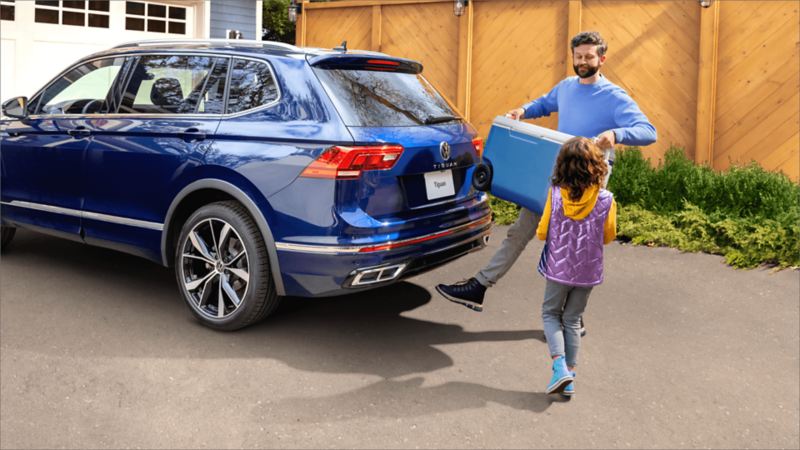A man holding large cooler standing beside his daughter puts his foot under the back of 2022 Tiguan to activate the Easy Open  tailgate. 