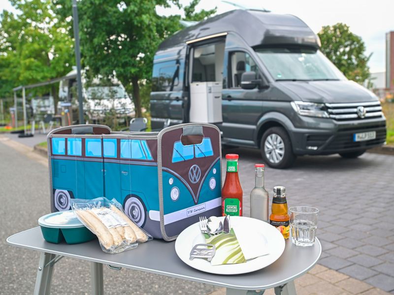 Auf einem Tischen stehen Utensilien fürs Grillen, sowie eine VW Tasche im Bulli Design und im Hintergrund parken ein VW Gran California und VW California.