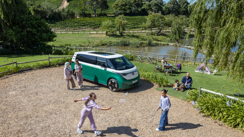 Van VW garé dans la nature près d’un lac, personnes jouant au badminton et en pique-nique