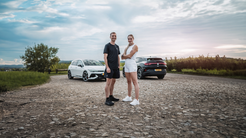  Sur un chemin de gravier, deux athlètes, un homme et une femme, posent devant deux voitures Volkswagen. À gauche se trouve une Volkswagen Golf R blanche, et à droite une Volkswagen ID.4 noire. L'arrière-plan est un paysage rural avec des champs, des arbres et un ciel nuageux au coucher du soleil